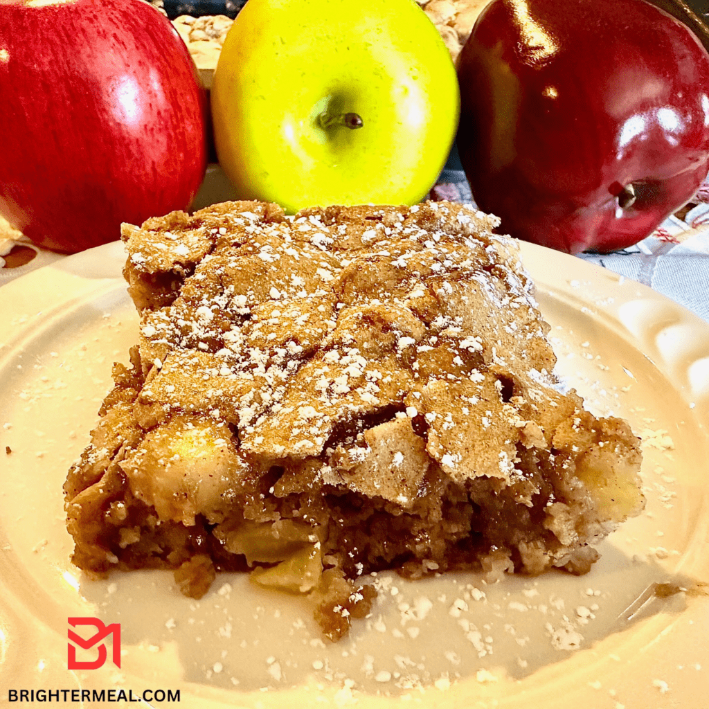 A tempting image of a freshly baked Apple Cake, adorned with a golden-brown crust and topped with a dusting of powdered sugar. The rich aroma of cinnamon and baked apples wafts through the air, inviting you to indulge in this delectable dessert. The cake sits on a vintage plate, surrounded by a sprinkle of cinnamon sticks and slices of fresh apples, adding a touch of rustic charm to the sweet masterpiece.